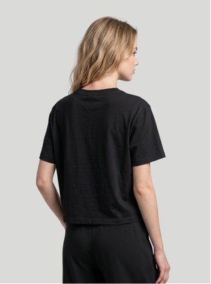 A woman with blonde hair is shown from the back wearing a black short-sleeve crop top and black pants against a solid light gray background.