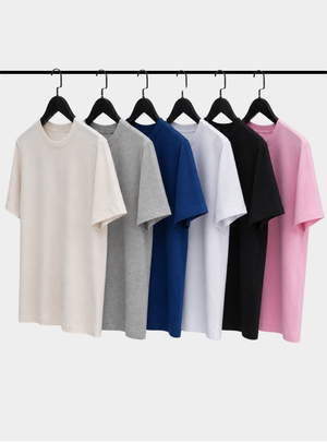 Six plain short-sleeve crew-neck T-shirts hanging on black hangers from a horizontal rack, shown in cream, gray, blue, white, black, and pink against a light gray background.
